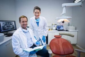 Two people in lab coats are sitting next to a dentist 's chair.